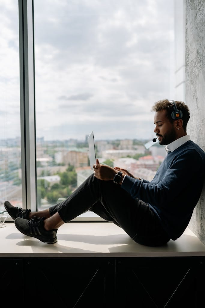 African American man working on laptop with headset by a large window. Urban view in background.