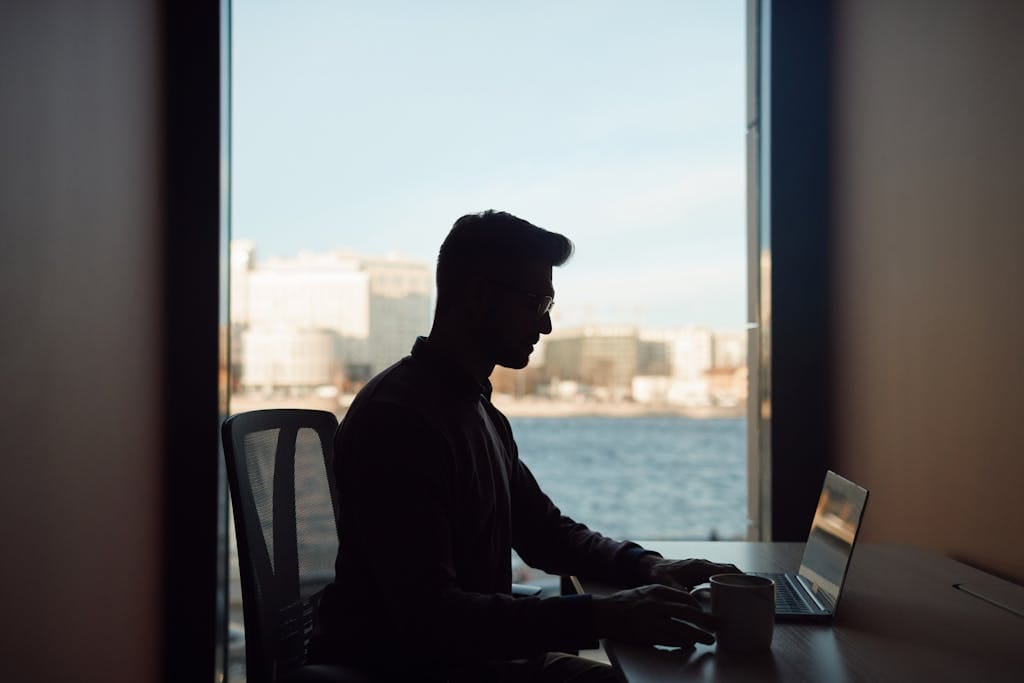 Silhouette of a man working on his laptop by a window overlooking a waterfront cityscape.